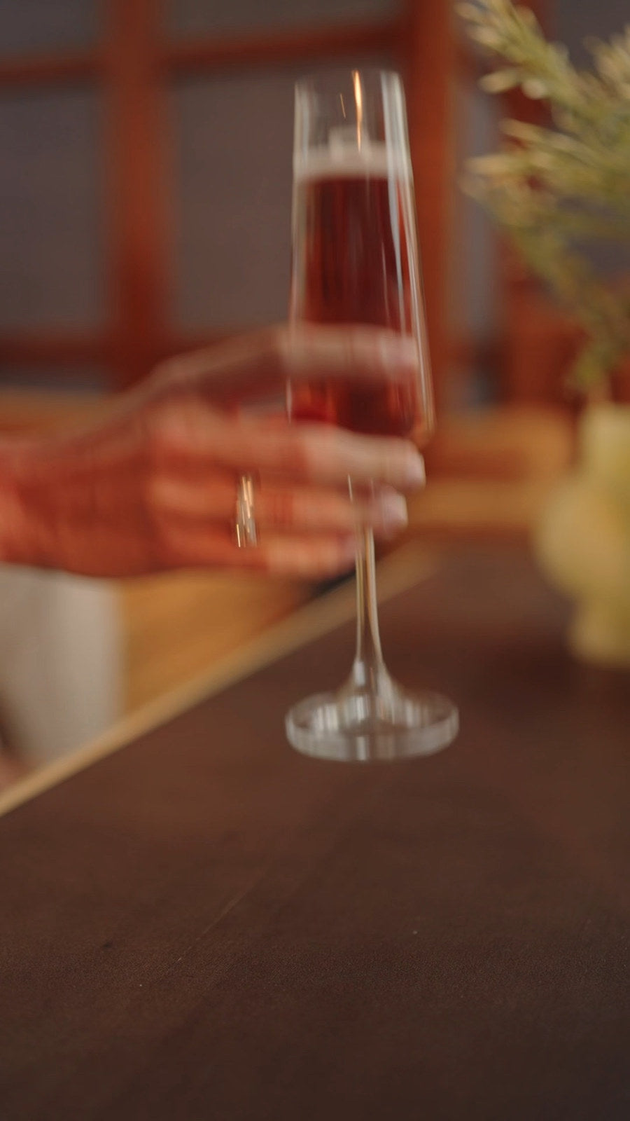 Film still from Chapter Two: her hand lifts a stemmed glass across a dark table, a ring on her finger.