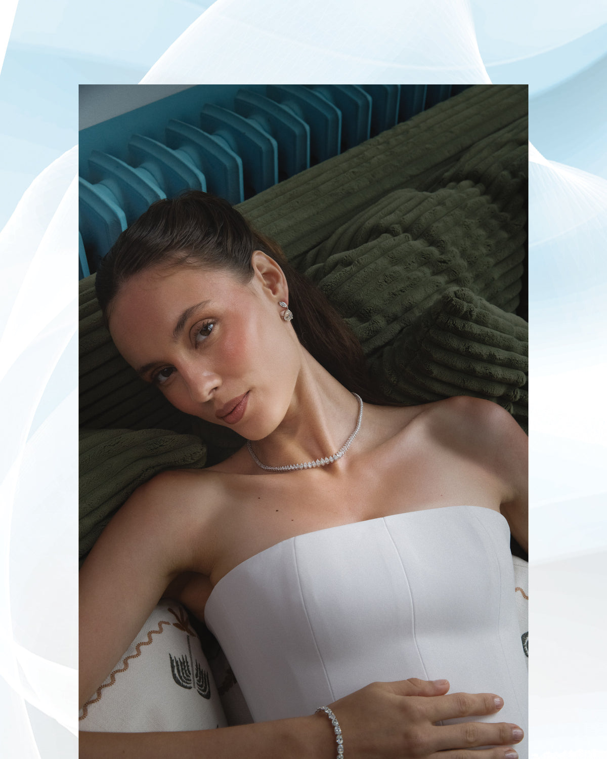 She lies back on a green sofa in a white dress, necklace and bracelet on.